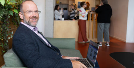 Smiling man in a blazer sits on a green couch using a laptop in a lobby; front desk and people in the background.