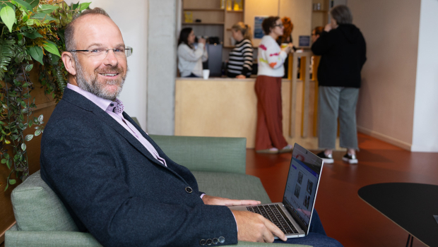 Smiling man in a blazer sits on a green couch using a laptop in a lobby; front desk and people in the background.